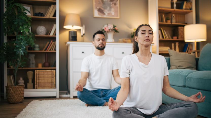 Picture of a man and a woman sitting on the floor in a modern apartment and practicing yoga. They wear white shirts.