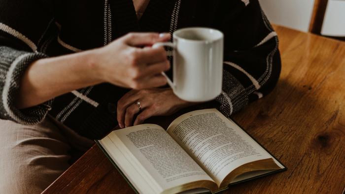 A photo showing a cropped image of a person reading a book at a wooden table and holding a white mug
