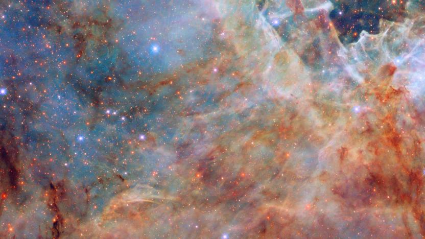 An image of the outskirts of the Tarantula Nebula showing "A section of a nebula, made up of layers of coloured clouds of gas, of varying thickness. In the background are bluish, translucent and wispy clouds; on top of these are stretches of redder and darker, clumpy dust, mostly along the bottom and right. In the bottom left corner are some dense bars of dust that block light and appear black. Small stars are scattered across the nebula."