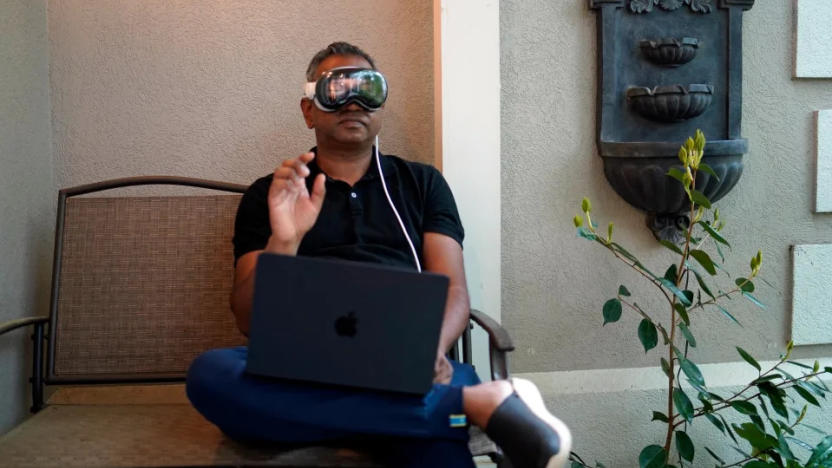 Engadget editor Devindra Hardawar is pictured sitting on an outdoor bench wearing the Apple Vision Pro headset and holding his hand up in a gesture in front of him