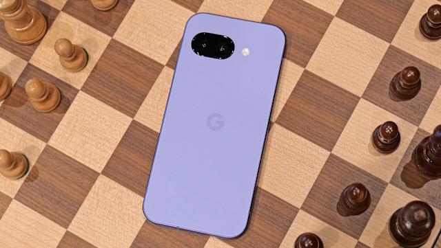 The purple Pixel 9a laying on the middle of a chess board, with black and white chess pieces on either side. The phone's screen is facing down.
