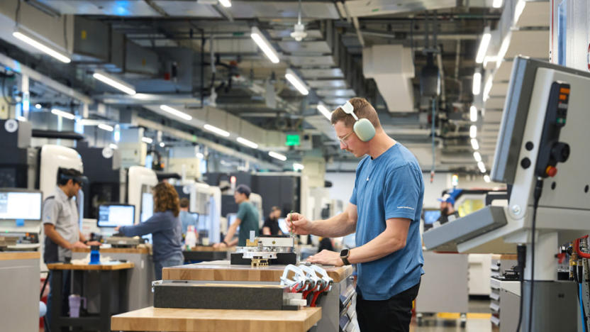 Groups of workers in an Apple manufacturing plant. A worker in the foreground wears AirPods Max headphones while working with a machine.