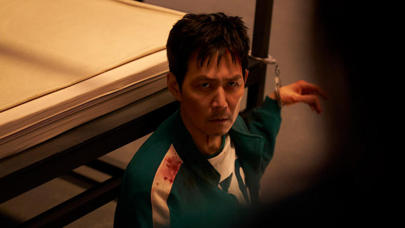 A man wearing a green tracksuit stained with blood is handcuffed to a bedframe.